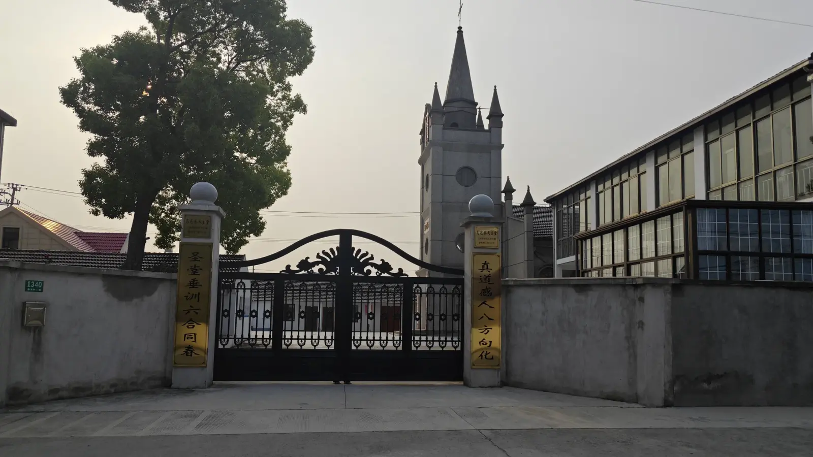 Ornate black iron gate with gold-lettered gateposts, a tree and a grey church tower beyond the wall.