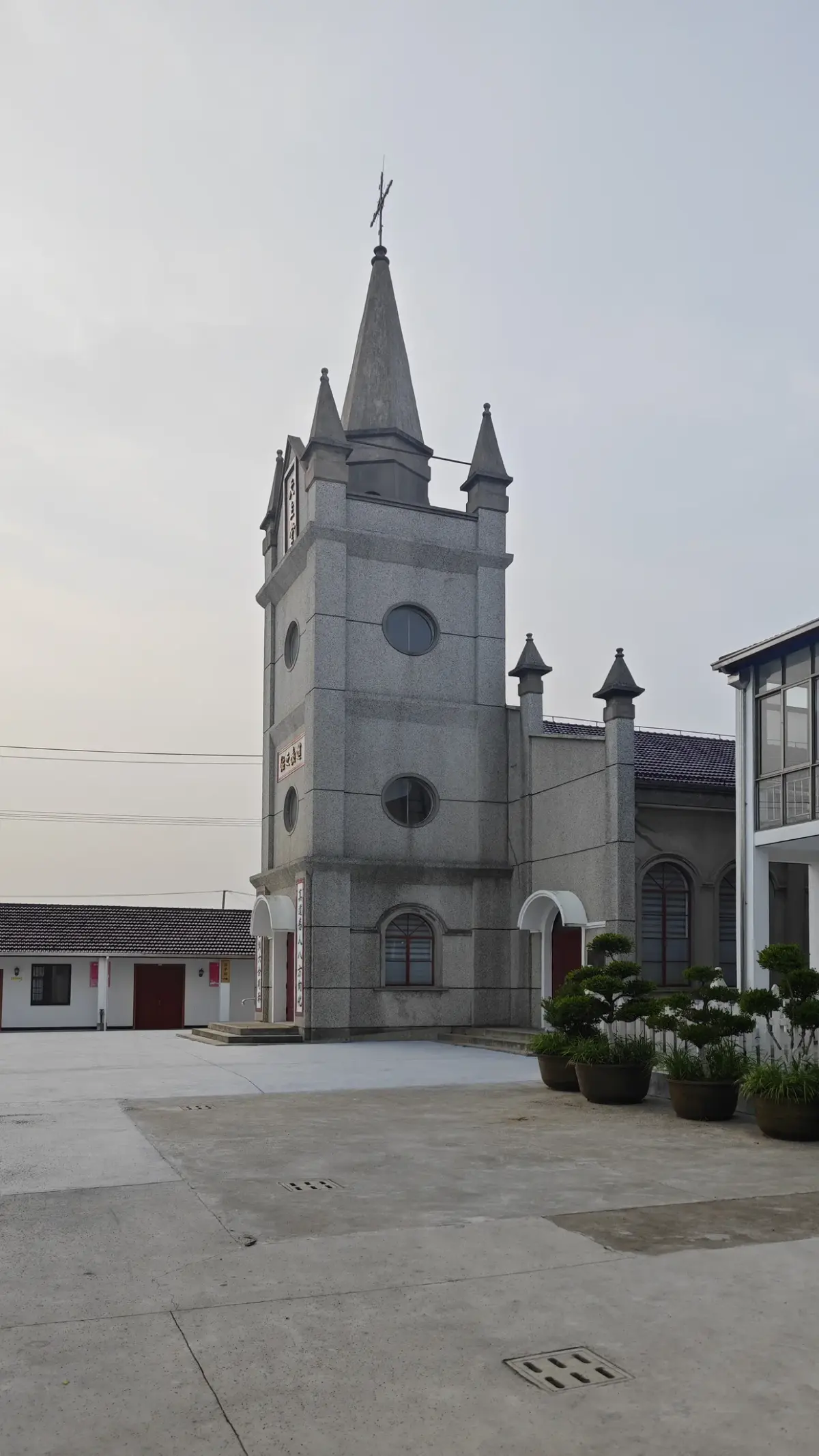 A grey bell tower with round clock windows and a slim spire rising above a courtyard, with a glazed building to the right.