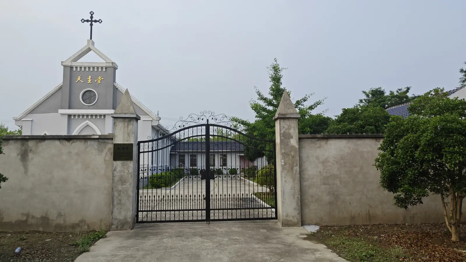 Church gable with a cross visible above a wall and a black iron gate with stone posts.