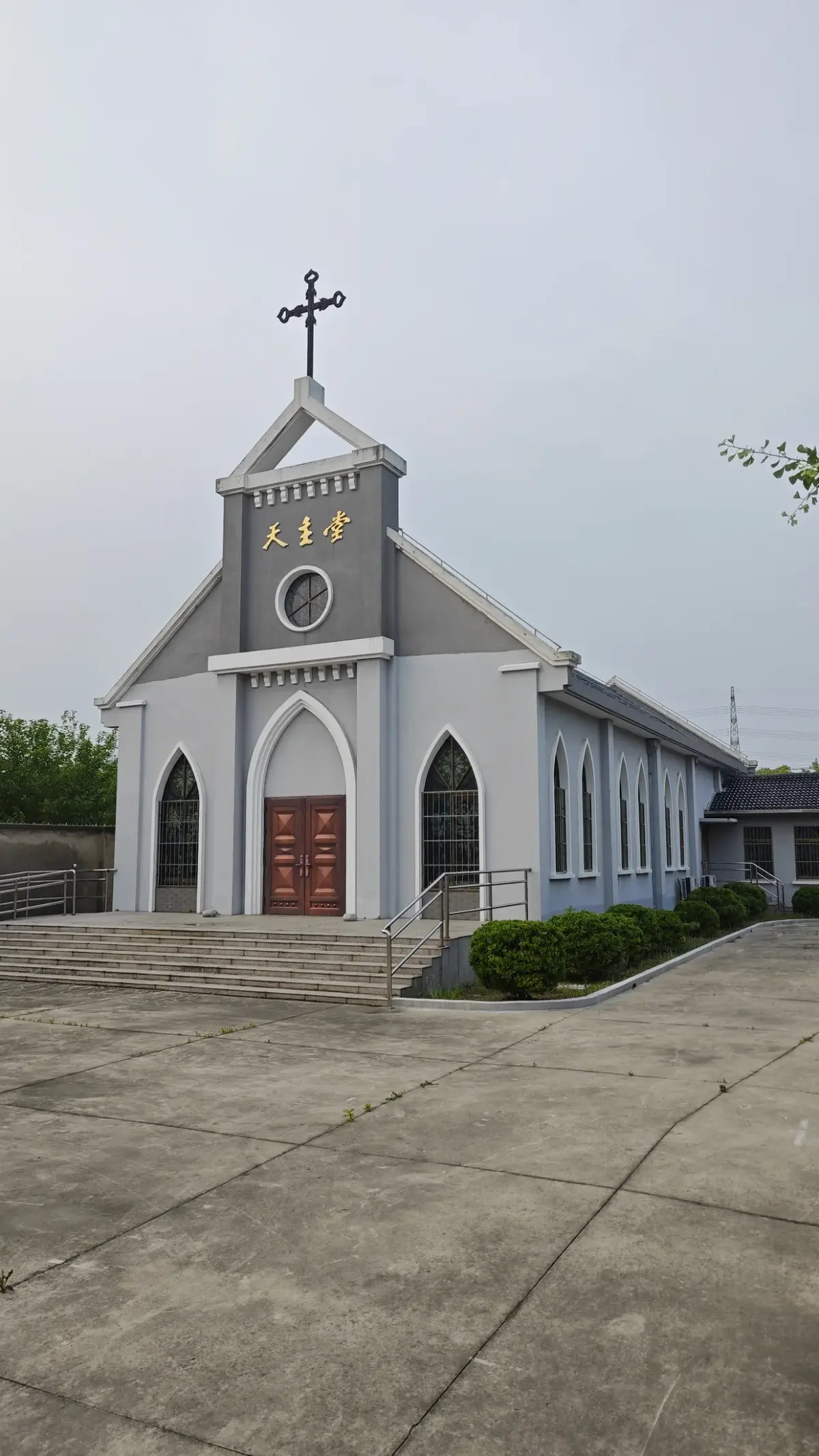 Portrait view of a grey and white gabled church front with a cross, pointed arched red doors and steps.