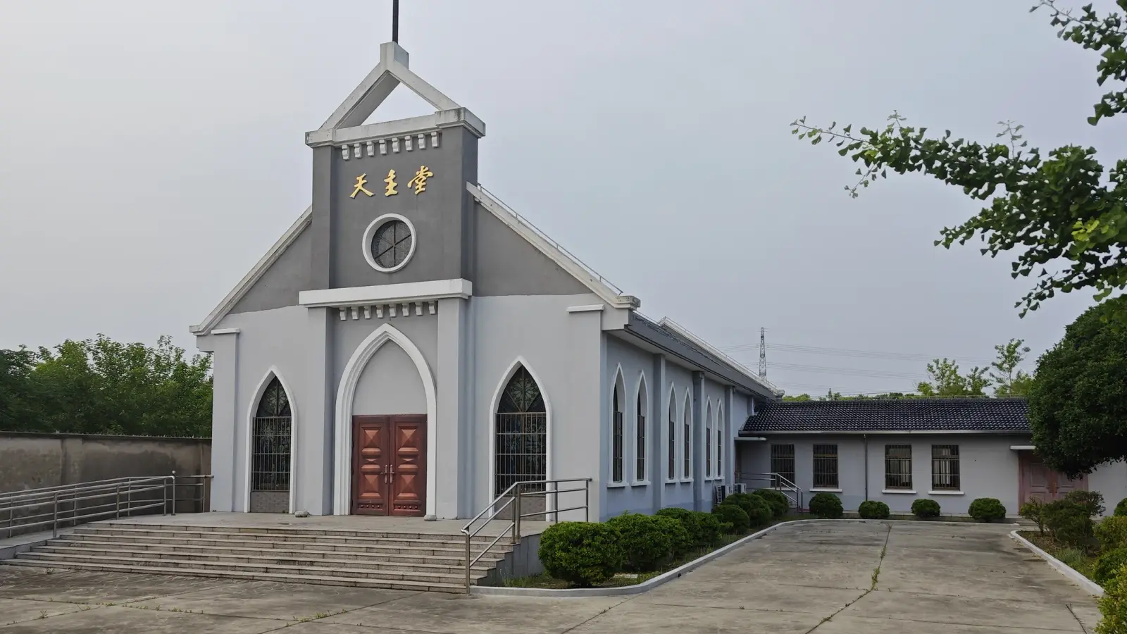 A grey and white church with a cross on the gable, red double doors, steps and a long low nave beside a paved yard.
