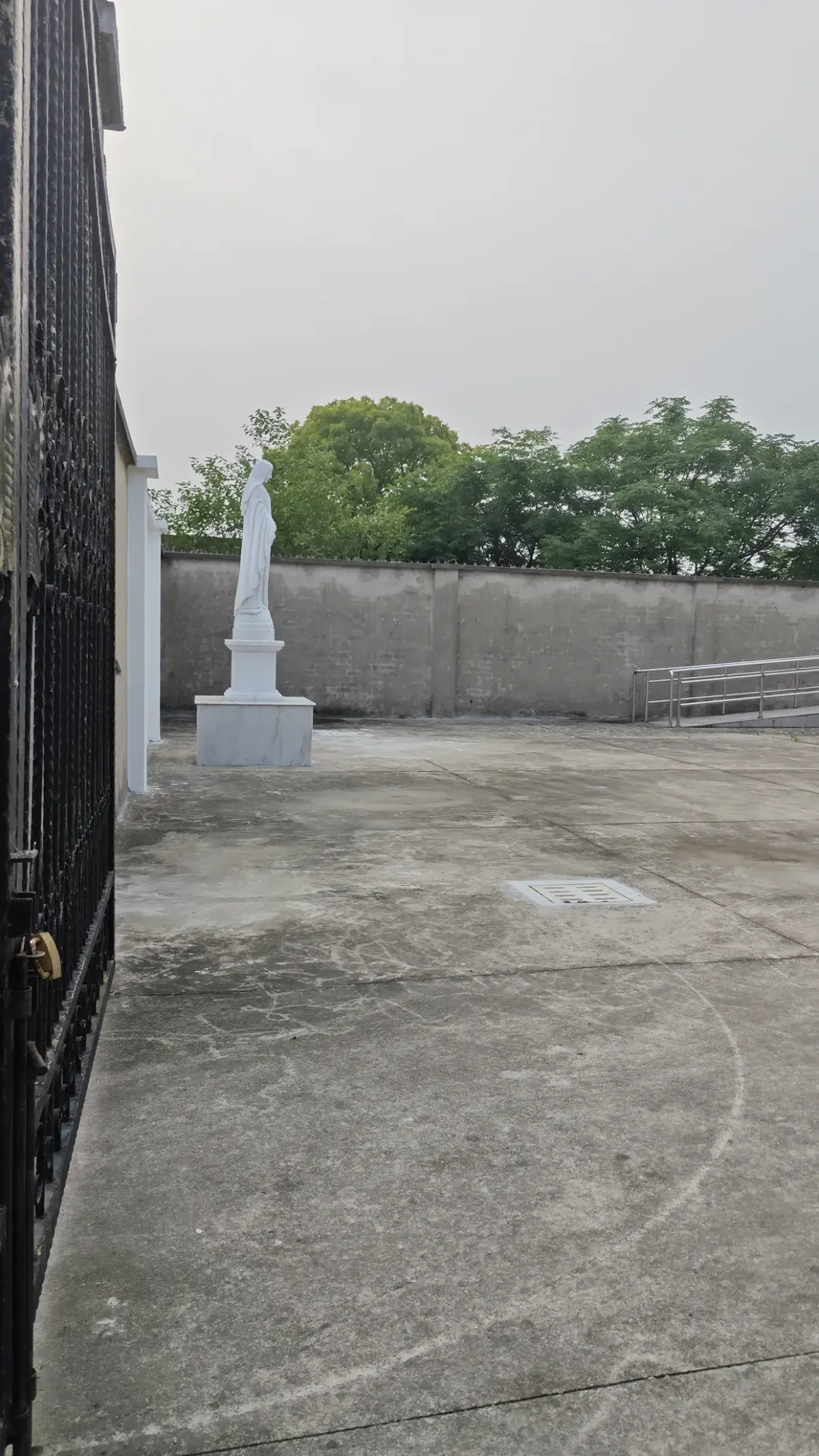 White statue standing against a wall in a paved churchyard, seen past gate bars.