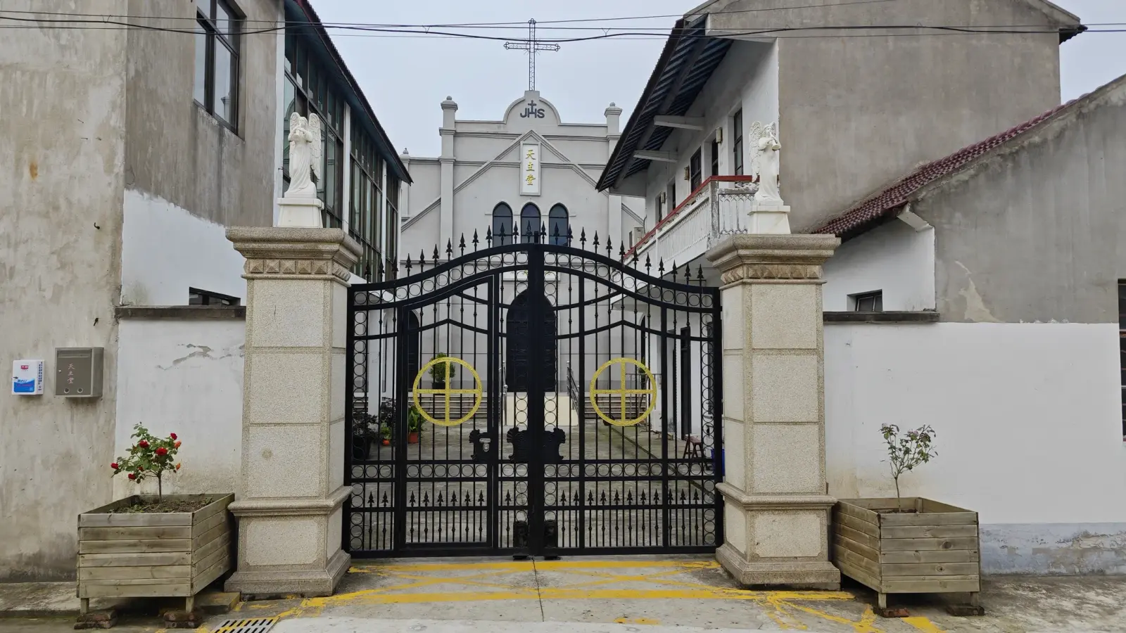 Black wrought-iron gate between stone gateposts topped with statues, and a church gable with a cross beyond.