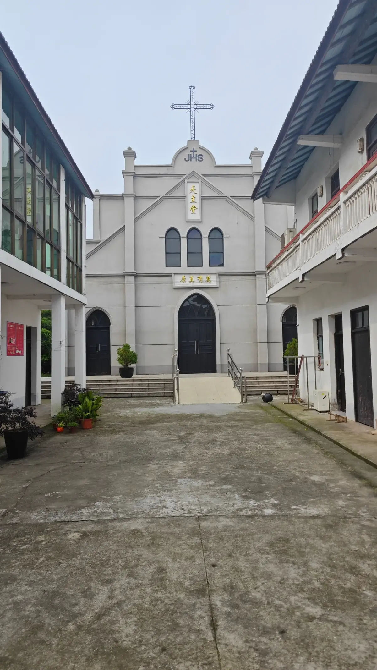 Courtyard view toward a pale grey church front with a cross, three lancet windows and a pointed doorway, flanked by two-storey buildings.