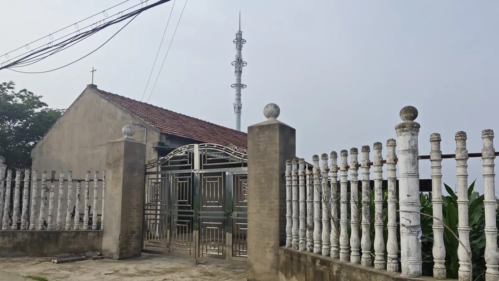 Plain gabled rural chapel with a tiled roof behind an ornate metal gate and white balustrade fence.
