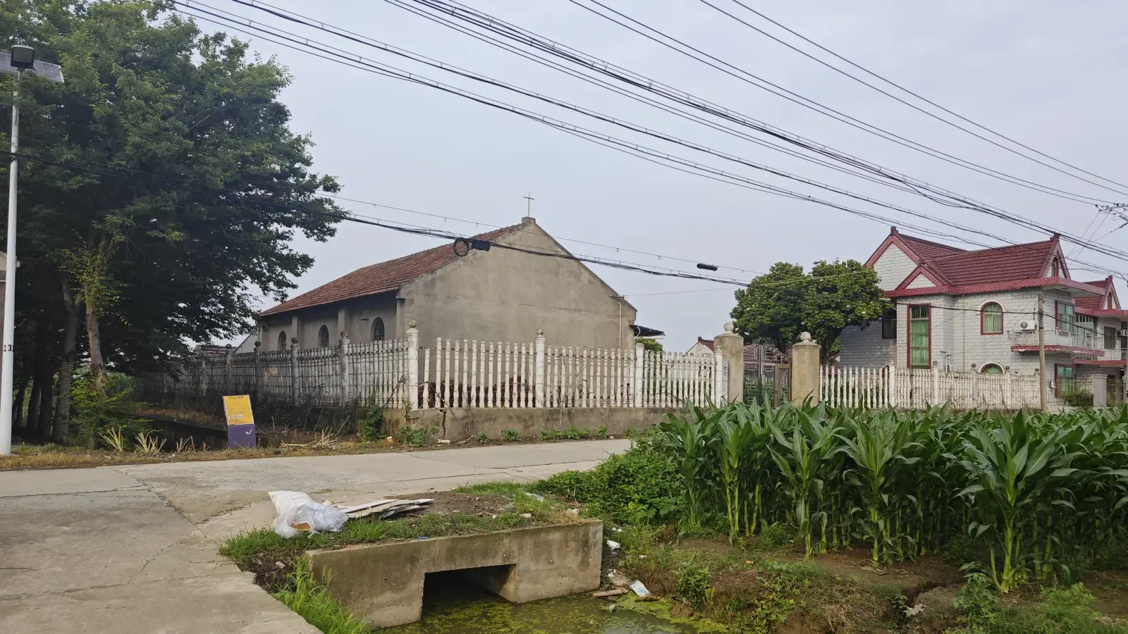 Distant view of a plain gabled chapel beside a lane, maize and village houses.