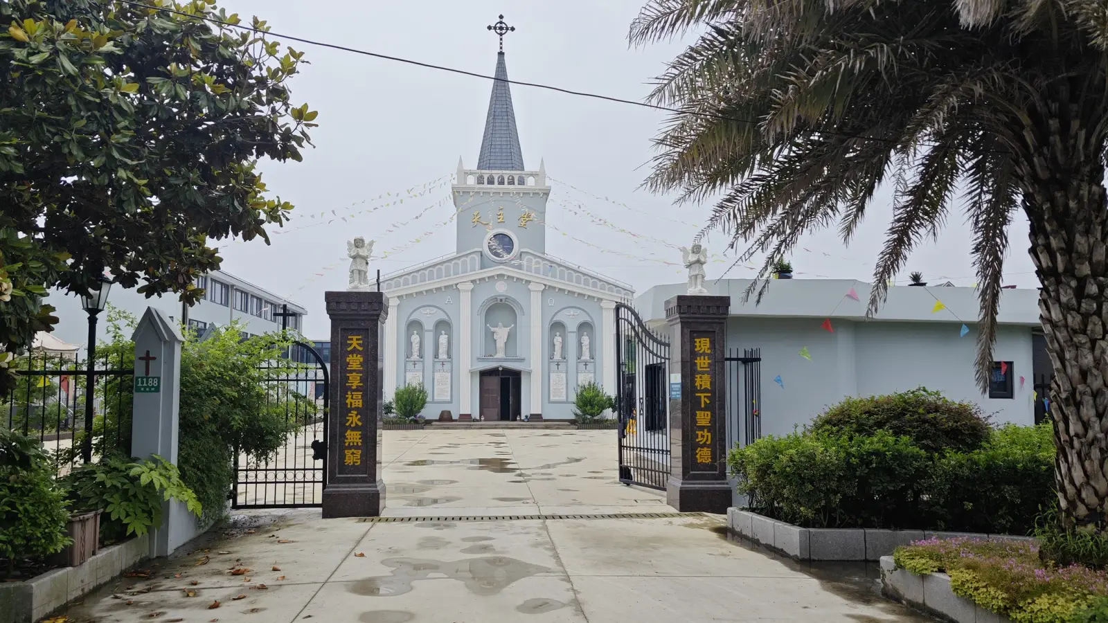 Drive leading to a blue-grey church with a spire, between gateposts with gold lettering and a palm tree.