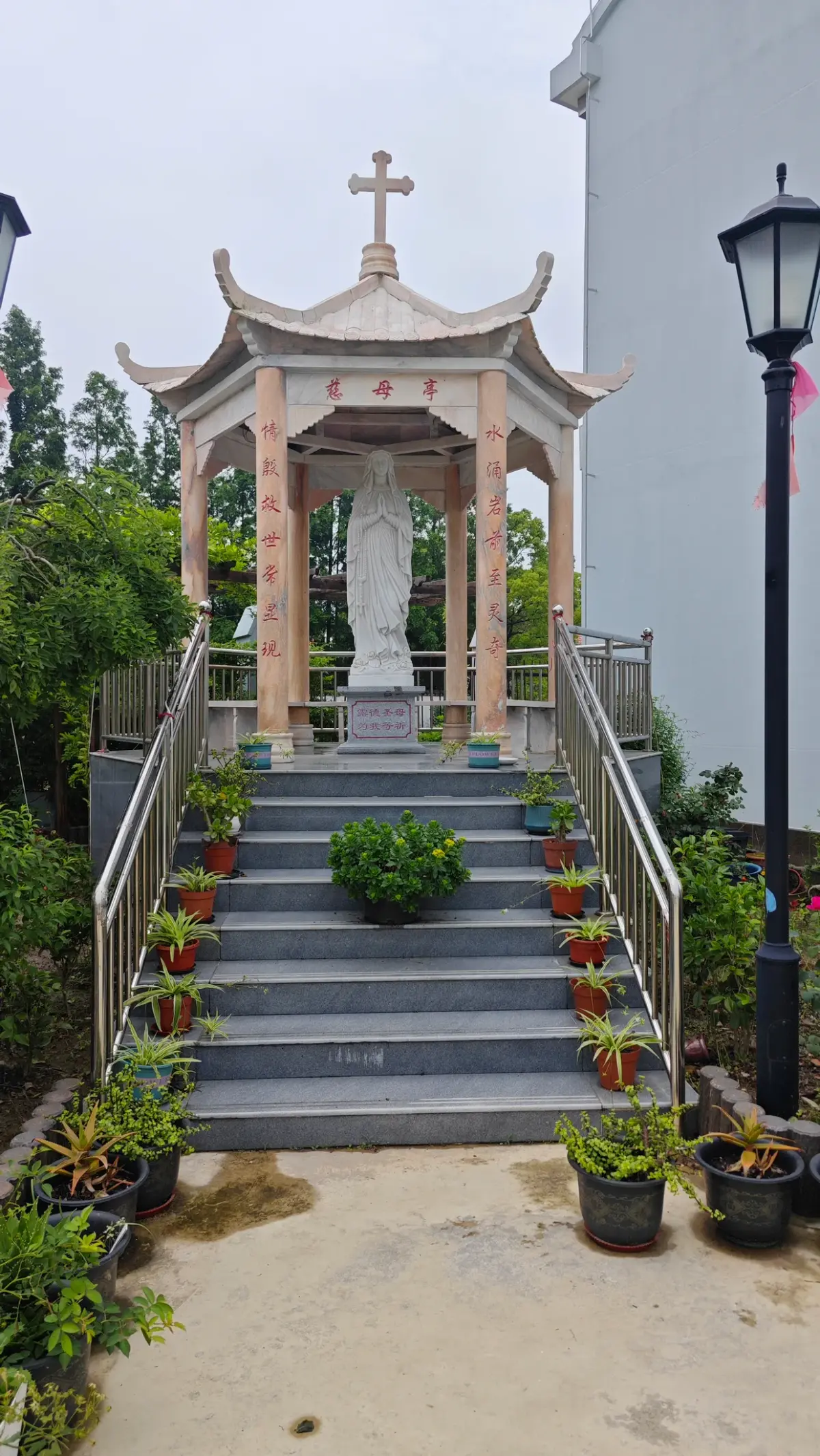 White statue of the Virgin Mary under a pavilion with a cross, at the top of a stepped path with potted plants.