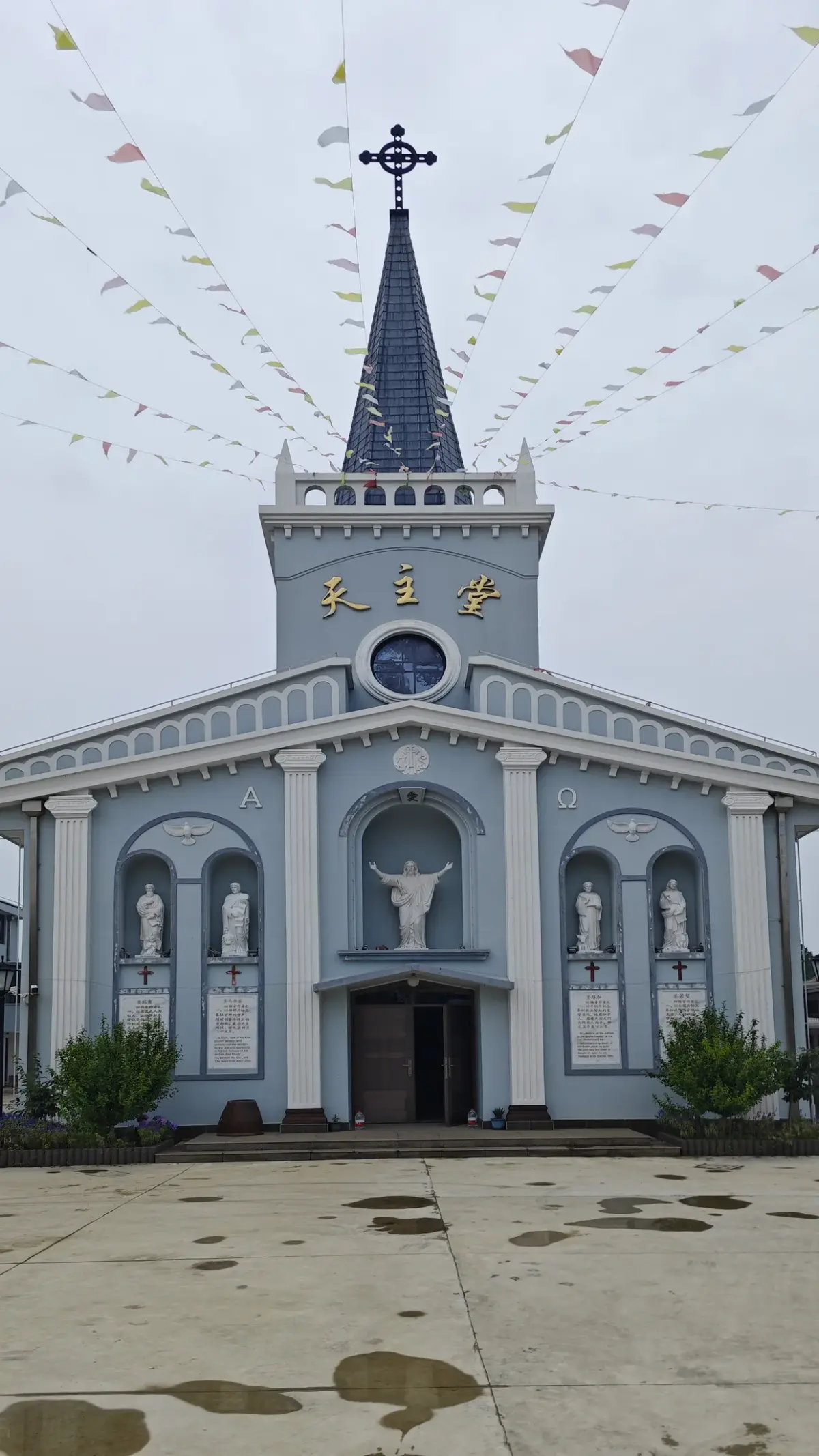Frontal view of a blue-grey church with a pointed spire and clock, a central Christ statue niche, side niches and a Chinese name board.