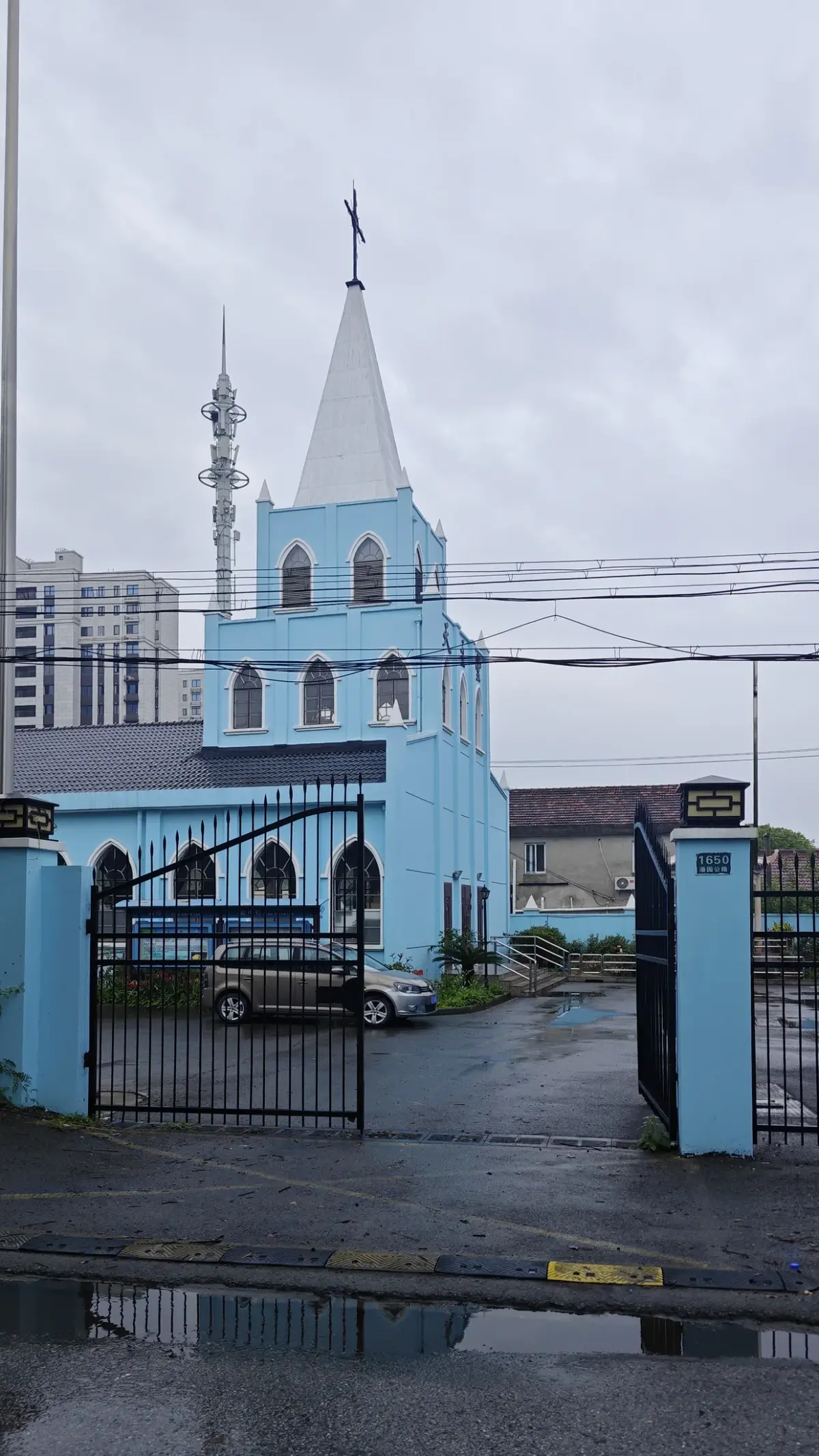 Blue church tower and white spire seen through an open iron gate, with the street number 1650 on the gatepost.