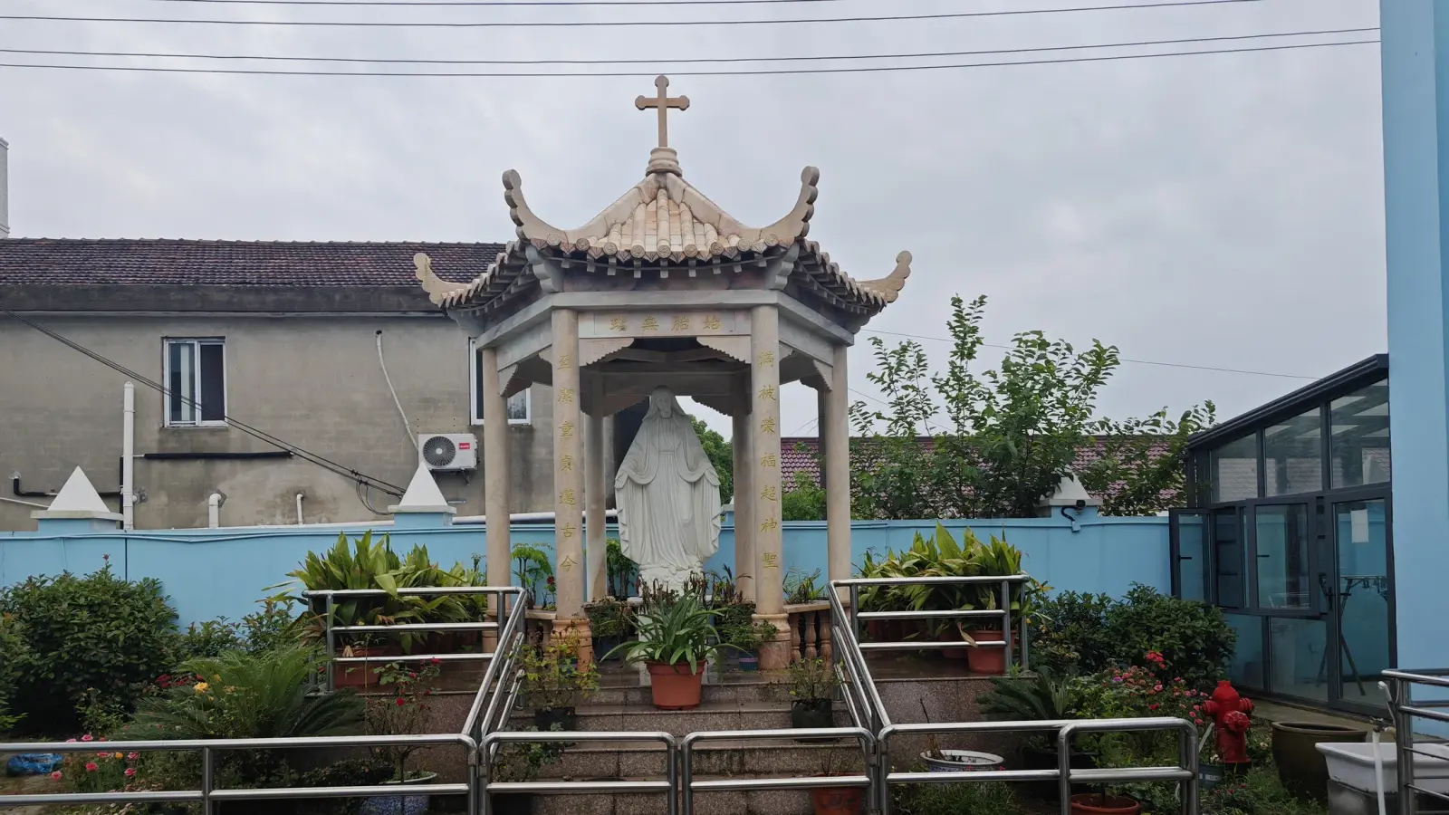 A statue of the Virgin Mary beneath a hexagonal pavilion with a cross on top, ringed by plants.