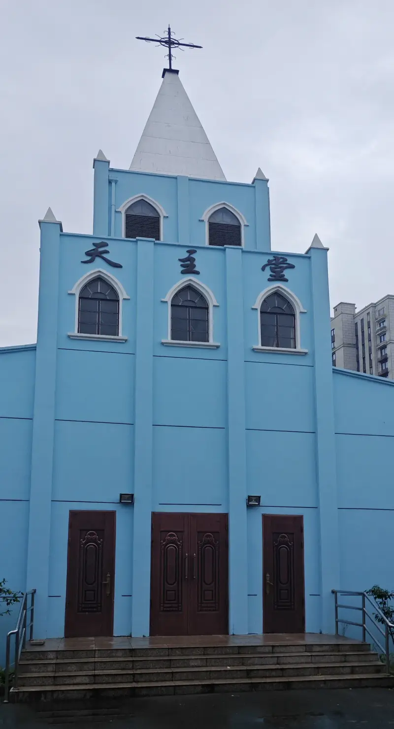 Frontal view of a pale-blue church facade with a white spire, pointed windows, a Chinese-character name board and three wooden doors.